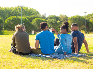 Back view of multiethnic friends gathering in park. Rear view of happy young male and female friends sitting on green lawn in park. Weekend concept