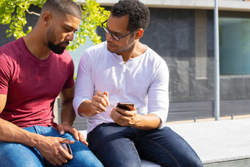 Serious mix raced man explaining how app working to his male friend. Two men in casual sitting on parapet outside, using phone and discussing content. Digital technology concept