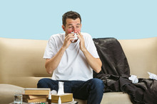 Young Man Suffering From Hausehold Dust Or Seasonal Allergy. Sneezing In The Napkin And Sitting Surrounded By Used Napkins On The Floor And Sofa. Taking Medicines With No Result. Healthcare Concept.