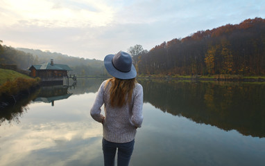 Stylish young woman in a warm sweater and hat, standing on pier by the lake in the park at autumn. Back view.