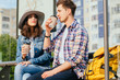 © Iryna - Elegant young brunette woman in hat and denim jacket and her boyfriend drinking coffee outdoor while waitig for bus or tram on stop with urban cityscape on background.