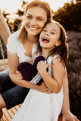 Wall Mural - portrait of a mother and her girl embracing and sitting on wooden swing looking at camera against su