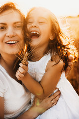 Close up portrait of lovely daughter and her mother embracing and laughing against sunset in a wheat field.