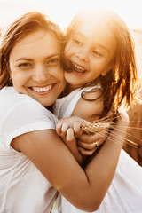 Wall Mural - portrait of a lovely mother embracing her daughter laughing against sunset dressed in white.