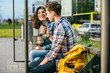 © Iryna - Positive relax couple traveler drinking coffee outdoor at stop while waiting tram, sits on bench at sunny morning.