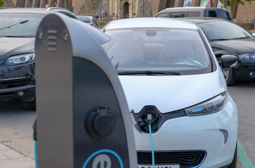 Close up of white Electric car at charging station with power cable supply plugged. Eco-friendly alternative energy concept. Power supply connect to electric vehicle. Front view. Chip electric cars.