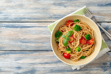 Wall Mural - Whole wheat spaghetti pasta with broccoli, cherry tomatoes and basil in bowl on wooden background. Top view. Copy space.