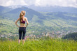 © bilanol - Back view of slim young woman standing on grassy valley on background of green mountains on sunny summer day.
