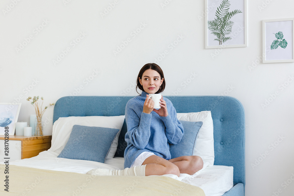 Girl in a blue sweater in interior Hygge style with a cup in hand sits on the bed - obrazy, fototapety, plakaty 