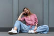 © Денис Бухлаев - young European girl, natural appearance, long hair, beautiful face, blue wide pants, crimson color shirt, white sneakers sit gray background. Linen womens clothing.