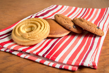 On the table on a red towel in striped cakes is to coffee: delicate cookies, fritters, and two oatmeal cookies.