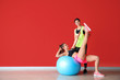 © Pixel-Shot - Young sporty woman with fitball doing exercises under supervision of her personal trainer near color wall