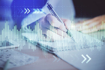 A woman hands writing information about stock market in notepad. Forex chart holograms in front. Concept of research. Multi exposure