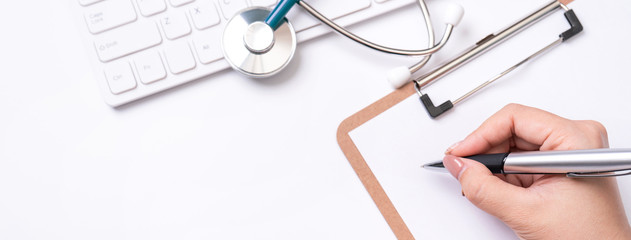 Wall Mural - Female doctor writing a medical record case over clipboard on white working table with stethoscope, computer keyboard. Top view, flat lay, copy space