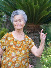 Senior Woman Stands Showing Hand Sign I Love You In The Garden