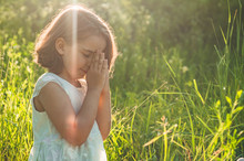 Little Girl Closed Her Eyes, Praying In A Field During Beautiful Sunset. Hands Folded In Prayer Concept For Faith, Spirituality And Religion. Peace, Hope, Dreams Concept