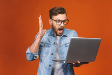 Surprised Amazed Young Business Man In Casual Standing And Holding Laptop Isolated On A Orange Background.