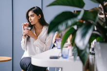Portrait Of A Beautiful, Attractive And Young Indian Asian Woman Sipping Her Iced Coffee As She Sits On Her Own In A Trendy Coffee Shop During The Day. She Is Wearing A White Blouse And Jeans.