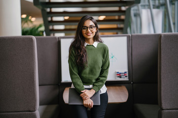 Portrait of a young, beautiful, intelligent and attractive Indian Asian MBA student smiling as she leans in a discussion booth in her campus. She is wearing a preppy outfit and smiling confidently.