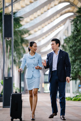 Wall Mural - Portrait of two well-dressed and professional Asian business people walking together in a modern city during the day. A Chinese man in a dark navy suit is talking to his female companion as they walk.