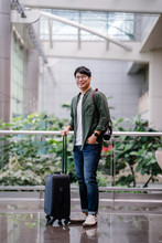 Portrait Of A Handsome, Young And Attractive Korean Asian Man Standing Next To His Travel Luggage In A Modern Airport. He Is A Tourist In A New Country And He Is Smiling Excitedly.
