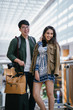 © Danon - A young interracial diverse couple casually dressed as they stand in the middle of an airport during the day. A young,handsome Korean man and his Indian woman companion are laughing out loud together.