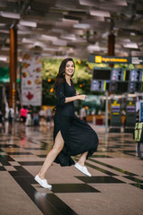 Wall Mural - Portrait of an attractive, young and athletic north Indian Asian woman smiling and laughing as she leaps and jumps indoors in a mall or airport. She is wearing a casual black flowing dress.