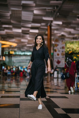 Wall Mural - Full body portrait of a young and attractive Indian Asian woman twirling in her casual and comfortable black dress and white sneakers. She is in a futuristic and modern airport or mall.