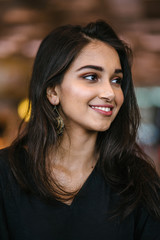 Wall Mural - A close up portrait of a beautiful, young, attractive and photogenic Indian Asian girl during the day in the city. She is wearing a casual black dress and smiling radiantly.