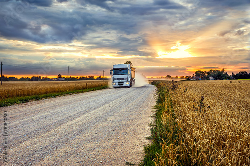 Grain truck on a rural road next to a rye field in the harvest season at sunset
