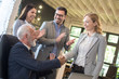© Bojan - Senior boss and middle aged female employee shaking hands with colleagues clapping hands around them in the office