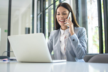 smiling businesswoman talking on mobile phone and using laptop in office