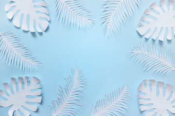 Handmade white paper cuts of tropical plant leaves and feathers arranged on blue background.