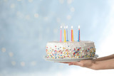 Fototapeta Tęcza - Woman holding birthday cake with burning candles against blurred background, closeup. Space for text