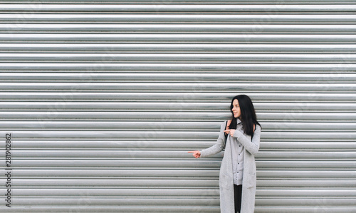Cuadros en Lienzo A young European girl in a gray cardigan stands against a metal textured wall and smiles, pointing her fingers to the left side of the background