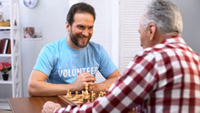 Smiling Middle Aged Male Volunteer Playing Chess With Senior Man In Nursing Home