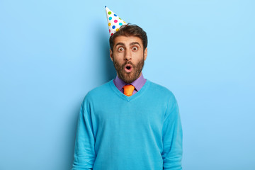 Portrait of shocked unshaven guy wears party hat, blue sweater with purple shirt and tie, surprised to be late on party, celebrates getting new job position with colleagues isolated on blue background