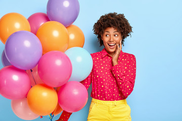 Wall Mural - Birthday greeting. Positive lady with Afro hairstyle, gazes aside, has party mood during happy holiday, wears fashionable bright clothing, holds bunch of colorful balloons, feels great pleasure