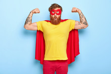 Emotive Surprised Male Hero Has Noble Qualities, Demonstrates Strength With Raised Arms, Has Strong Muscles, Dressed In Superhero Costume, Isolated On Blue, Has Strength To Face Dire Situation In Life