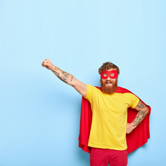 Brave cheerful man hero ready to fly, can sacrifice own life to help other, has extraordinary ability, wears red cape and mask, yellow t shirt, isolated on blue background, performs act of kindness