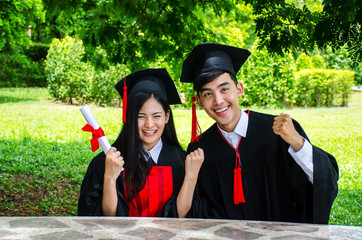 A man and woman couple dressed in black graduation gown or graduates with congratulations with hold diploma in hand is sitting..