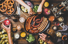 Flat-lay Of Octoberfest Dinner Table Concept With Grilled Sausages, Pretzel Pastry, Potatoes, Cucumber Salad, Sauces, Beers And Peoples Hands With Holding Snacks Over Dark Wooden Background, Top View