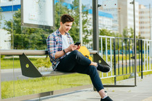 Male Student Wth Smart Phone In Hands Wearing Plaid Shirt Sitting On Bench While Waiting For Transport At Tram Or Bus Station.