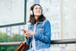 © Iryna - Smiling excited brunette woman wearing blue denim jacket with headphones looking away, relaxing, listening music while waiting at tram stop in a city.
