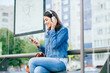 © Iryna - Smiling excited brunette woman wearing blue denim jacket with headphones looking away, relaxing, listening music while waiting at tram stop in a city.