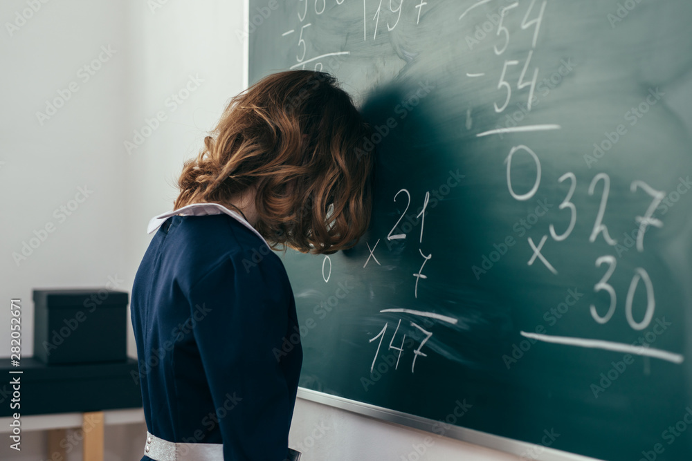 Sad girl pupil trying to solve an example. Schoolgirl stands with her forehead on the blackboard - obrazy, fototapety, plakaty 
