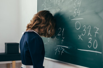 Wall Mural - Sad girl pupil trying to solve an example. Schoolgirl stands with her forehead on the blackboard
