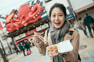 Wall Mural - takoyaki in styrofoam box on hands of young happy woman exciting in Osaka Japan. korean lady traveler trying japanese famous delicious snack in tsutenkaku street. cheerful female showing local food