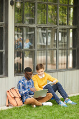 Wall Mural - Full length portrait of two cheerful international students studying together outdoors while sitting on green grass in college campus, copy space