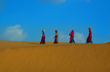 Indian women carrying heavy jugs of water on their head and walking on a yellow sand dune in the hot summer desert against blue sky.water crises, rajasthan, india